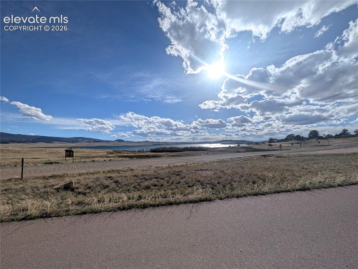 Paved road surface bordering a natural landscape with distant water, expansive sky, and scattered clouds