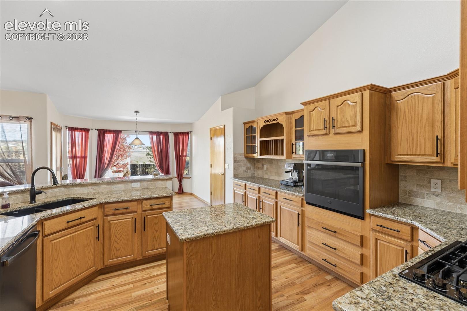 Kitchen featuring natural wood cabinetry, stone-finish countertops, built-in wall oven, central island, and vaulted ceilings