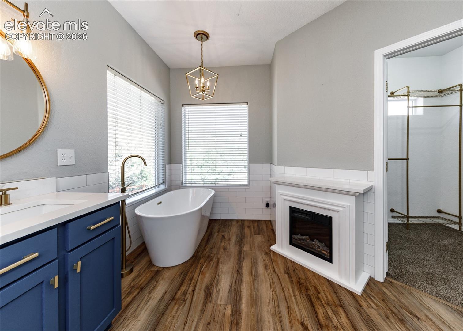 Freestanding soaking tub with a floor-mounted faucet, wood-finish flooring, white subway tile wainscoting, built-in electric fireplace, and blue vanity cabinetry with metallic hardware