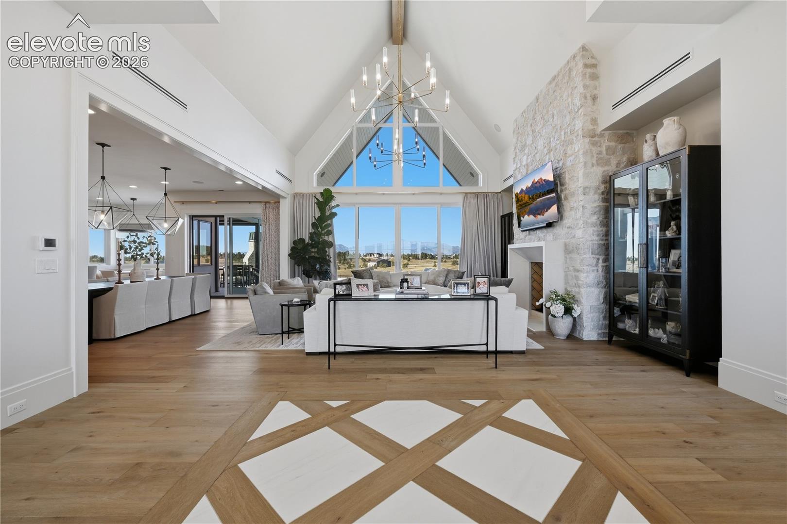 Vaulted ceiling with exposed structural beam, floor-to-ceiling stone fireplace, inlaid wood-finish flooring, and expansive geometric window wall
