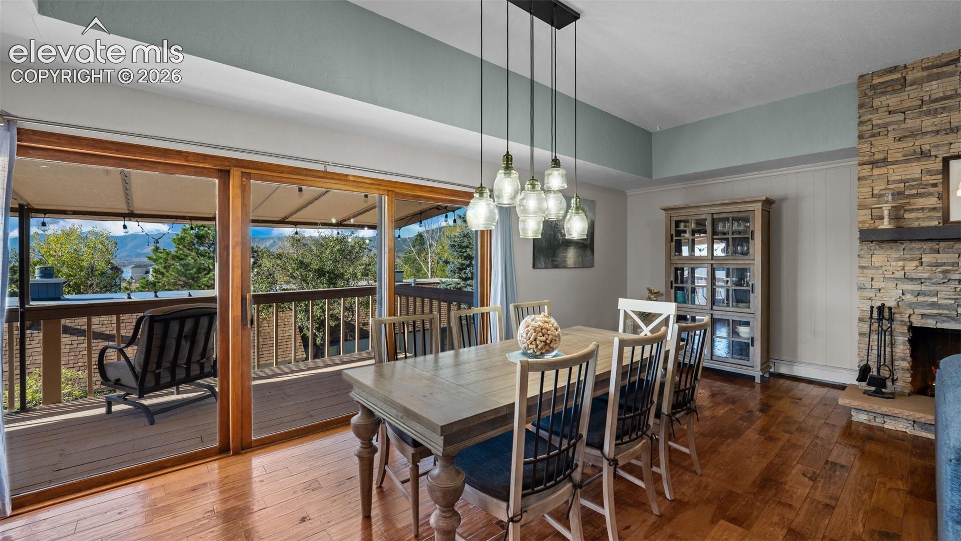 Dining area featuring wood-finish flooring, a floor-to-ceiling stone fireplace, multi-pendant light fixture, and sliding glass doors opening to a wood deck