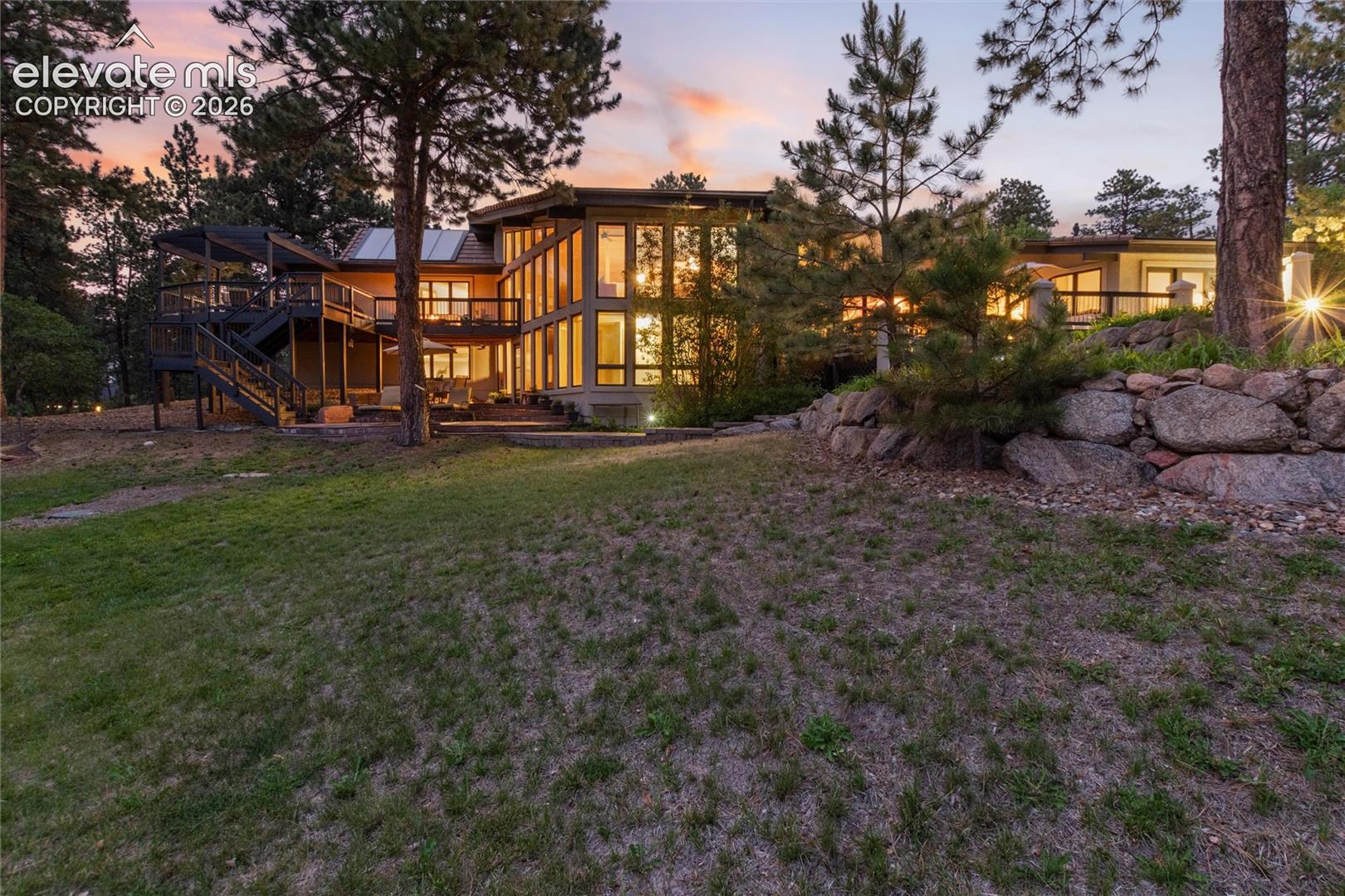 Contemporary multi-level residence featuring extensive window walls, multiple outdoor decks, a stone retaining wall, and mature trees