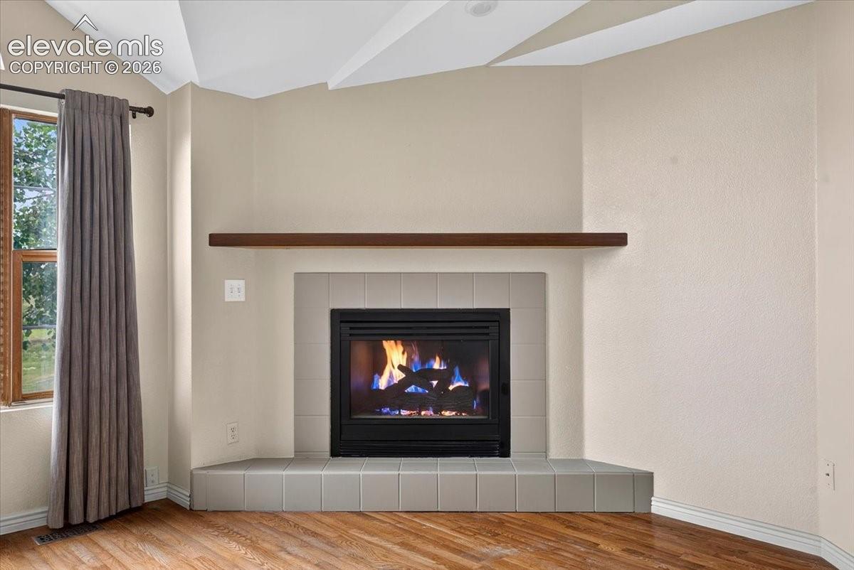 Corner fireplace with neutral square-tile surround, elevated tile hearth, wood-finish mantel, and vaulted ceiling