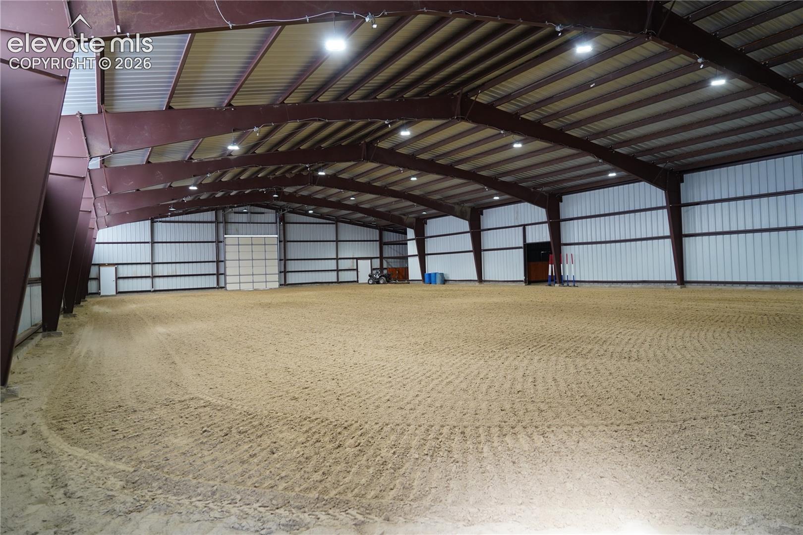 Expansive indoor arena with a sand footing, high ceilings with exposed steel beams, and multiple overhead light fixtures