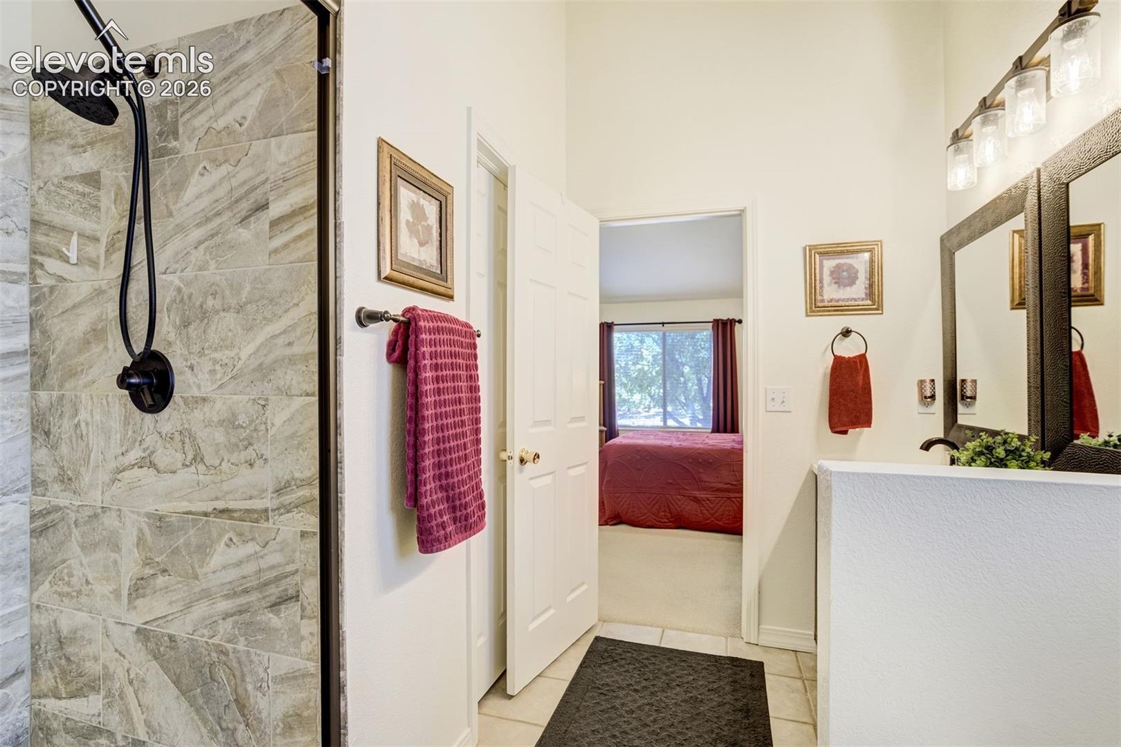 Primary Bathroom featuring a walk-in shower with stone-finish tile surround, a matte black rainfall showerhead, a dual-mirror vanity, and industrial-style multi-light fixture