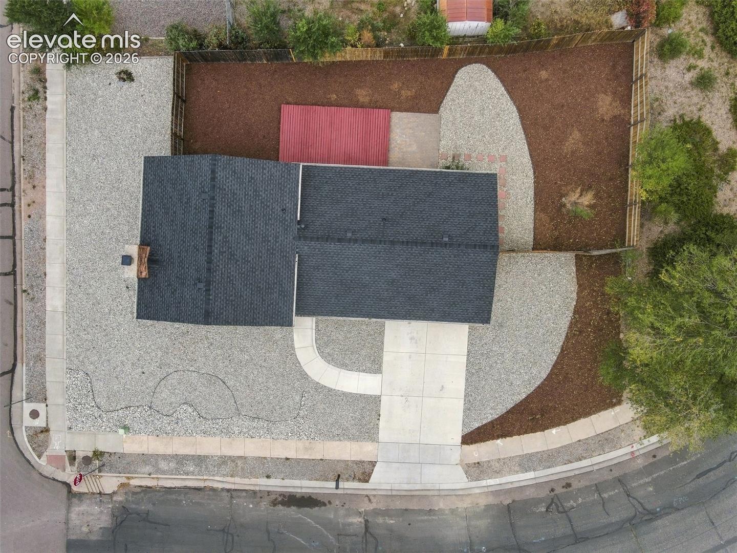 Aerial view of the property featuring a dark-shingled roof, a paved concrete driveway, a dedicated outdoor wooden deck, and a fenced perimeter with mixed gravel and mulch landscaping