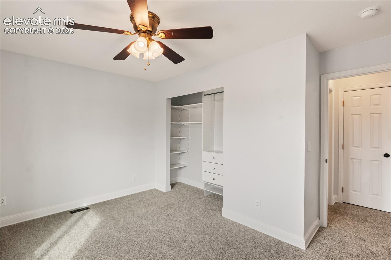 Neutral-toned carpeting, ceiling fan with integrated light fixture, and built-in closet featuring shelving and three-drawer storage