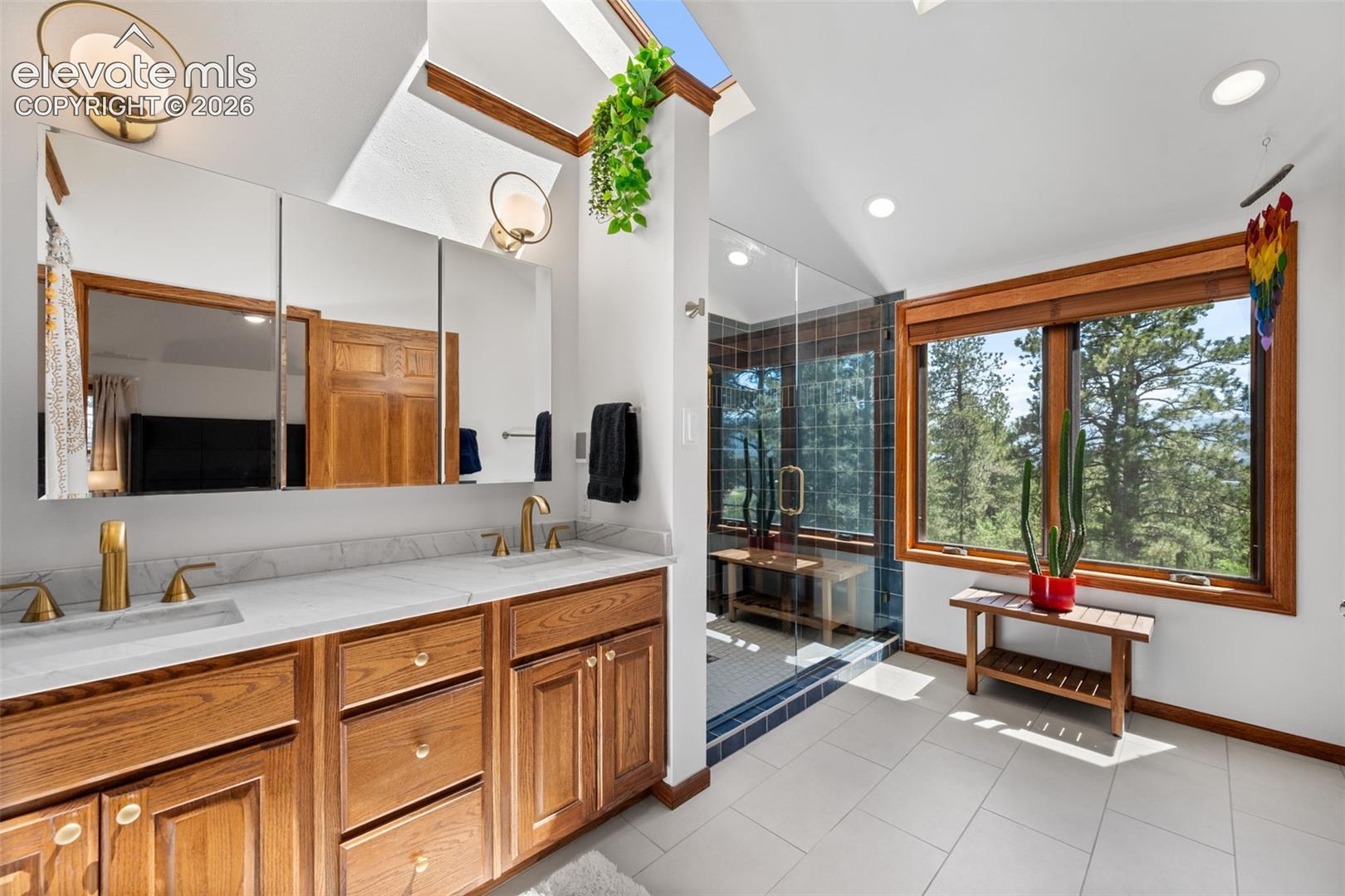 Bathroom featuring a wood-finish double vanity with dual gold-tone fixtures and white countertops, a glass-enclosed shower with dark tile, a skylight, and large windows with wood trim
