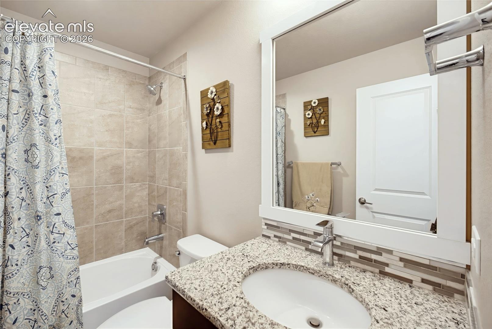 Bathroom featuring a speckled stone-finish countertop, undermount sink, multi-tone glass tile backsplash, framed wall mirror, and a tub-shower combination with floor-to-ceiling tile surround