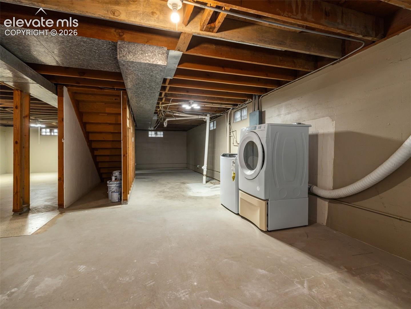 Unfinished basement with concrete flooring, exposed wood beam ceiling, full-size laundry appliances, and a built-in wood staircase