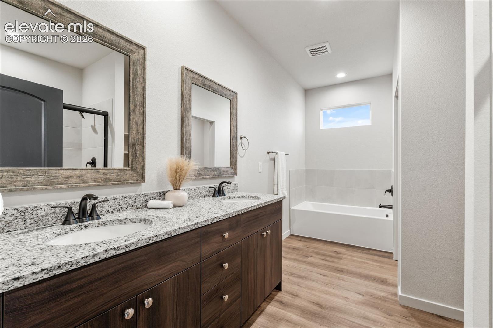 Dual-sink vanity with stone-finish countertop, dark wood-tone cabinetry, recessed lighting, and separate soaking tub