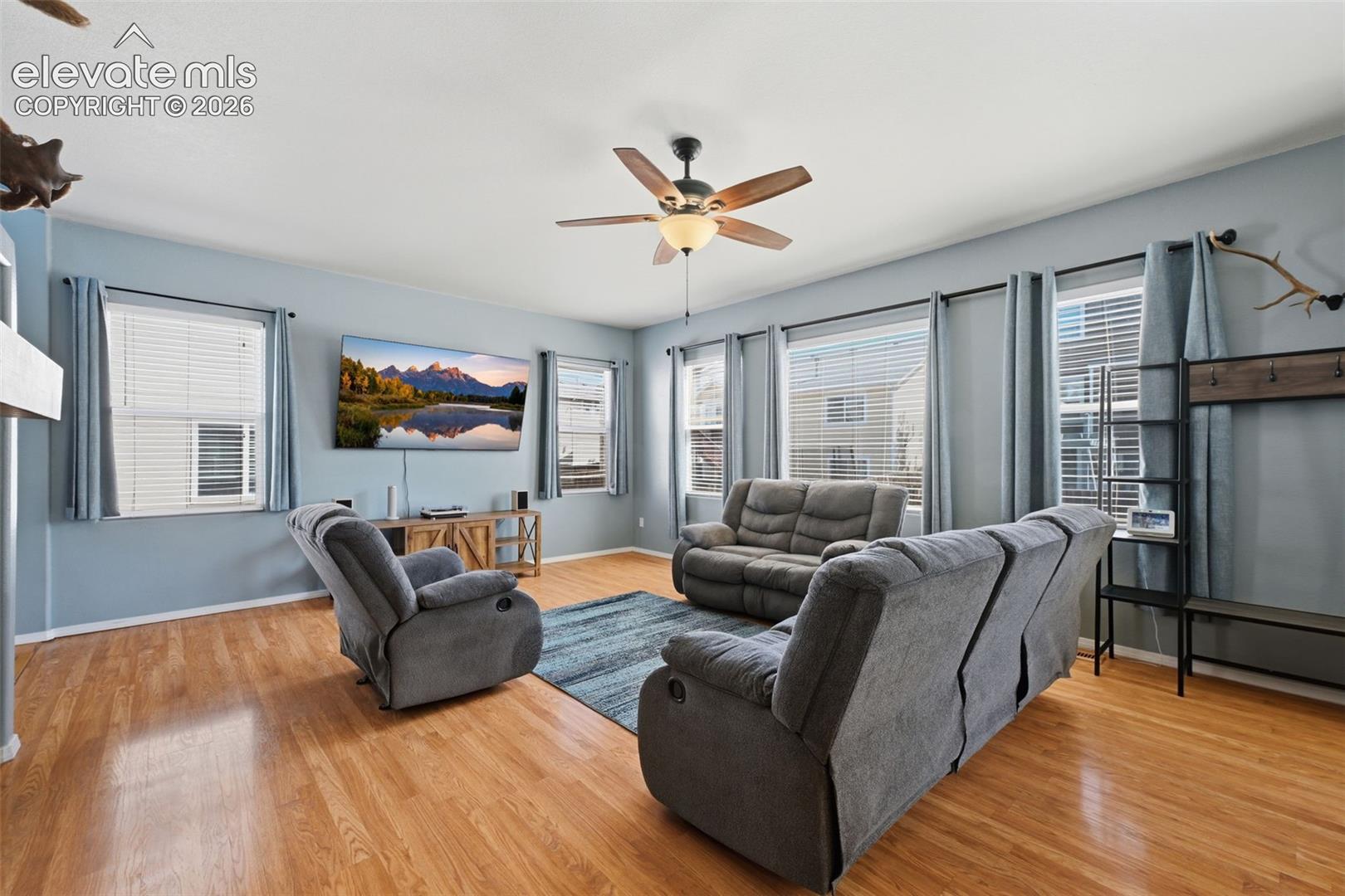 Hardwood-finish flooring, multiple windows with blinds, ceiling fan with integrated lighting, and a built-in wall unit