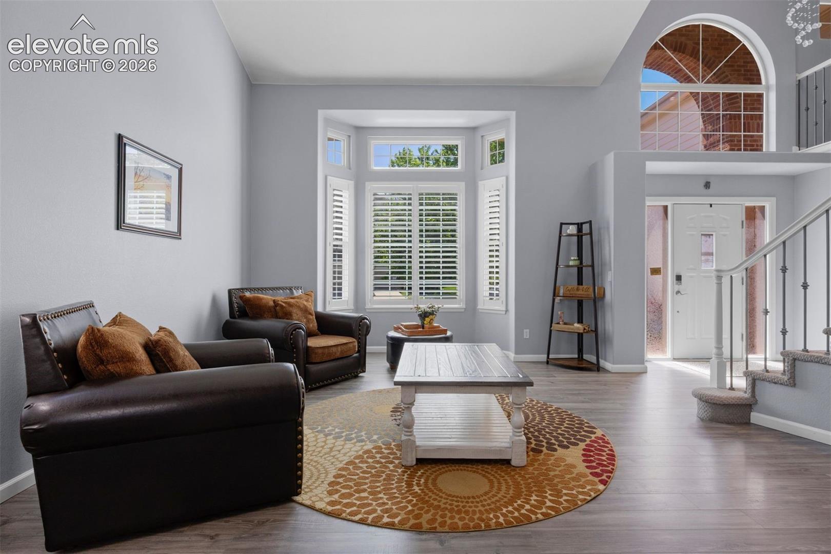 Living area featuring vinyl flooring, a bay window with plantation shutters, vaulted ceilings, and a staircase with balusters. Open and Bright