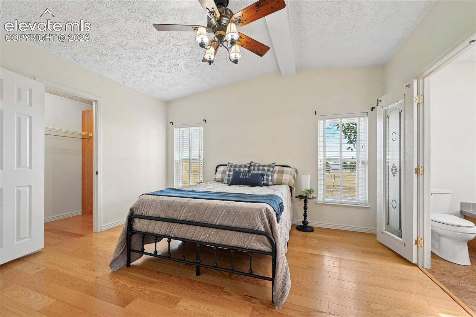 Vaulted ceiling with exposed beam, wood-finish flooring, two windows with blinds, and an attached closet with bi-fold doors
