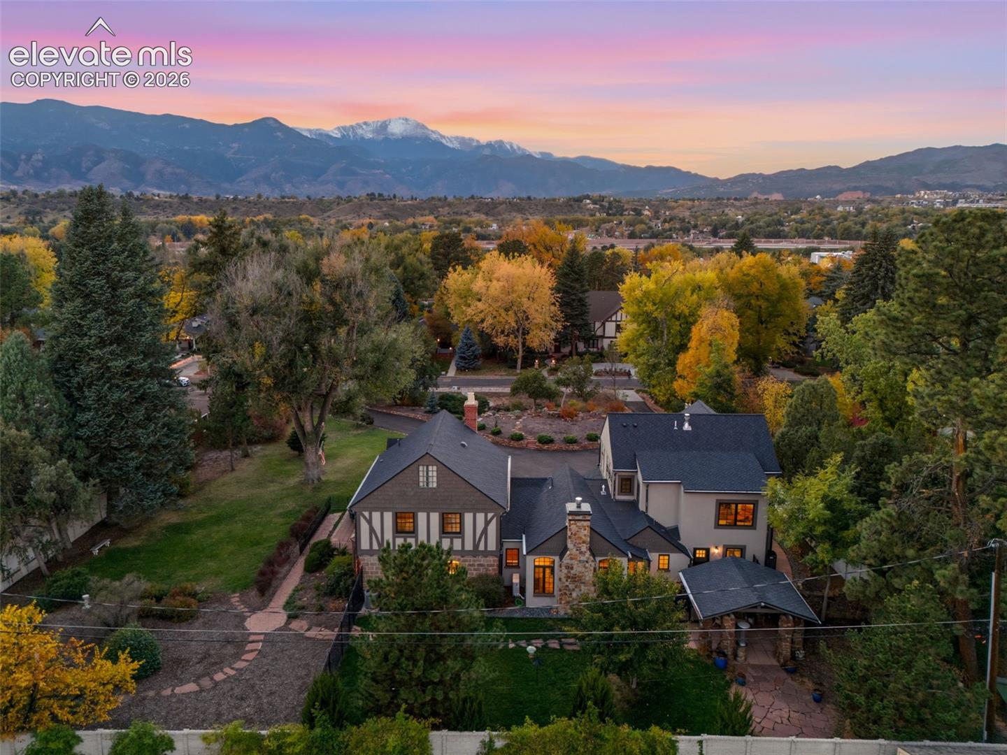 Tudor-style residence featuring stone and timber-frame exterior detailing, a multi-gabled shingle roof, stone masonry chimney, flagstone patio, and mature landscaping against a prominent mountain backdrop