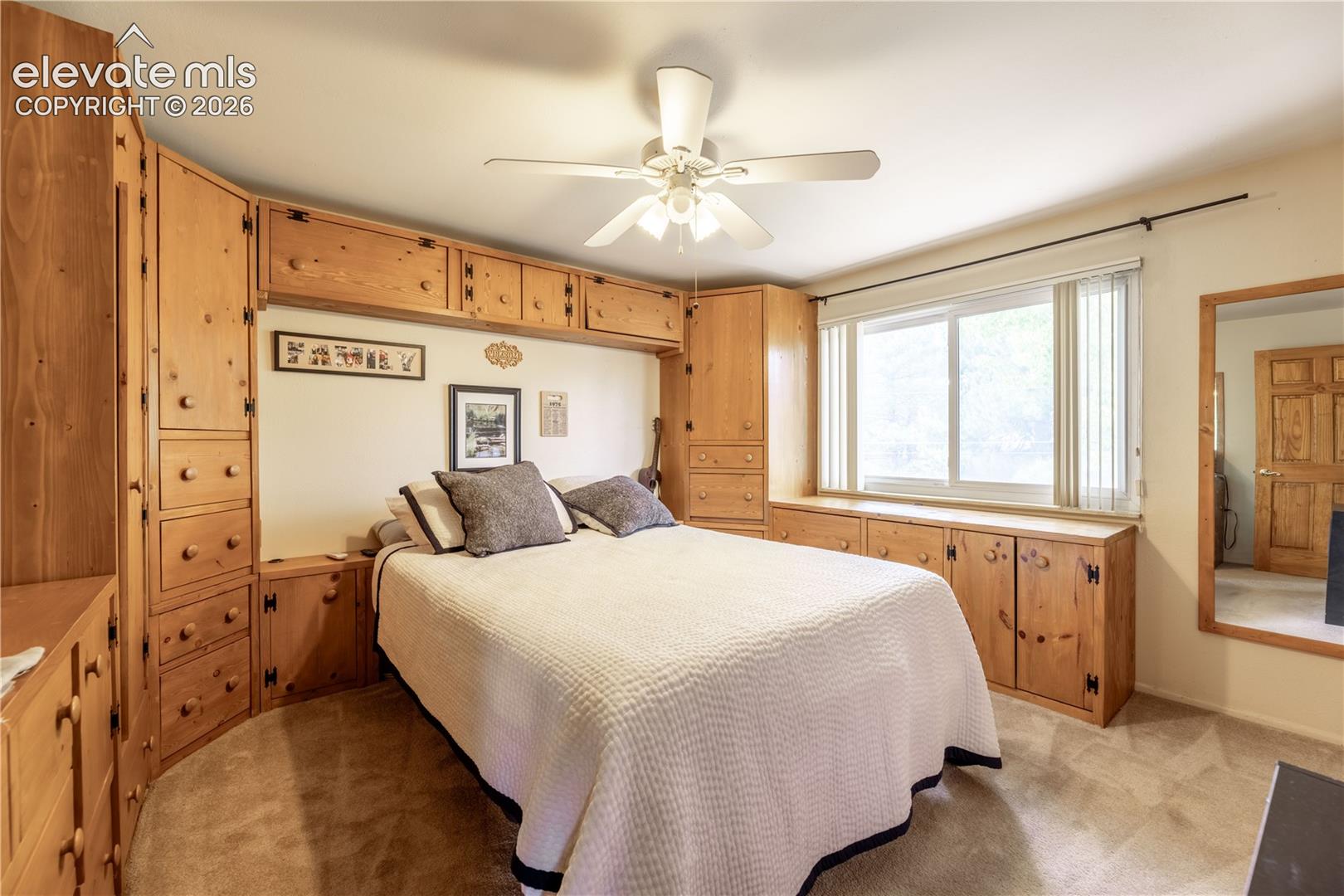 Custom built-in wood cabinetry spanning the wall, sliding window with vertical blinds, integrated ceiling fan with lighting, and wall-mounted mirror