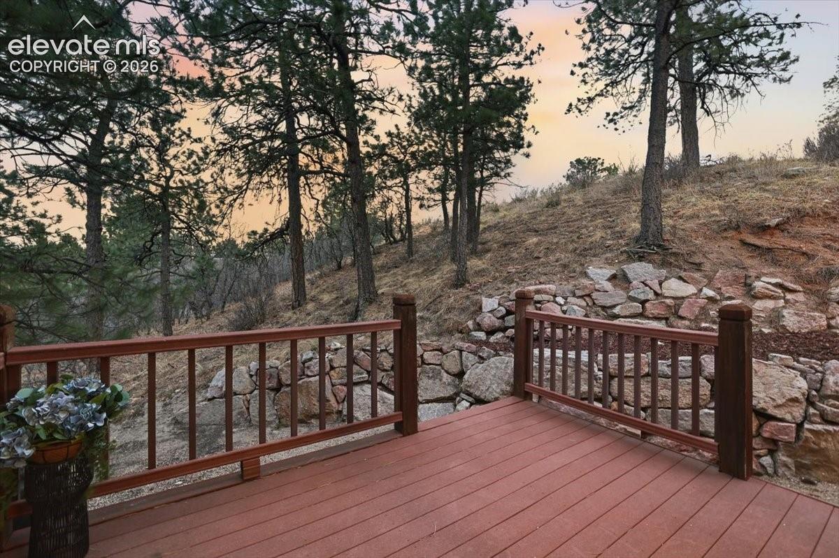 Expansive outdoor decking with wood-finish railing, set against a natural, sloped terrain featuring mature trees and a stone retaining wall