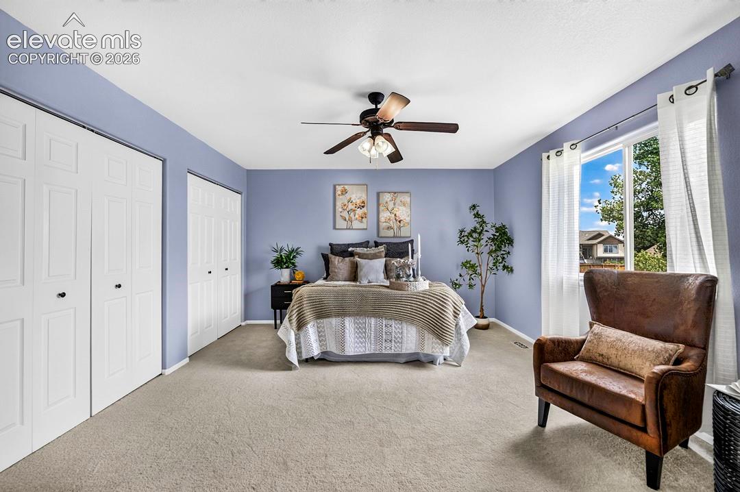 Room featuring neutral carpeting, dual white paneled closet doors, a multi-blade ceiling fan with integrated light fixture, and a window allowing natural light