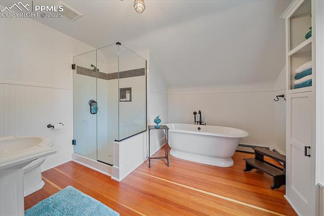 Full bath featuring wood finished floors, a freestanding bath, a stall shower, a wainscoted wall, and vaulted ceiling