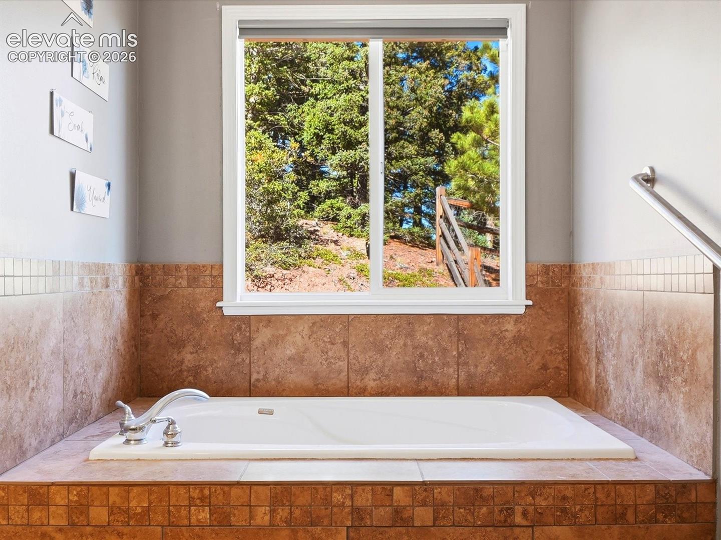 Built-in soaking tub with a tiled surround, chrome fixtures, and a window featuring a natural hillside backdrop