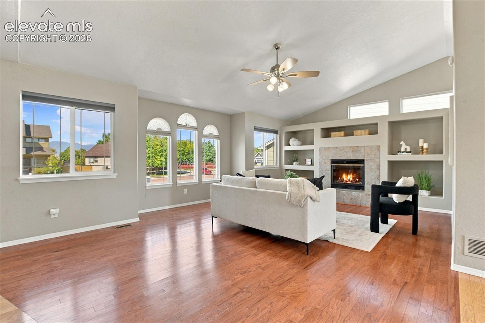 Vaulted ceiling with a ceiling fan, recessed built-in shelving, stone-surround fireplace, arched windows, and wood-finish flooring