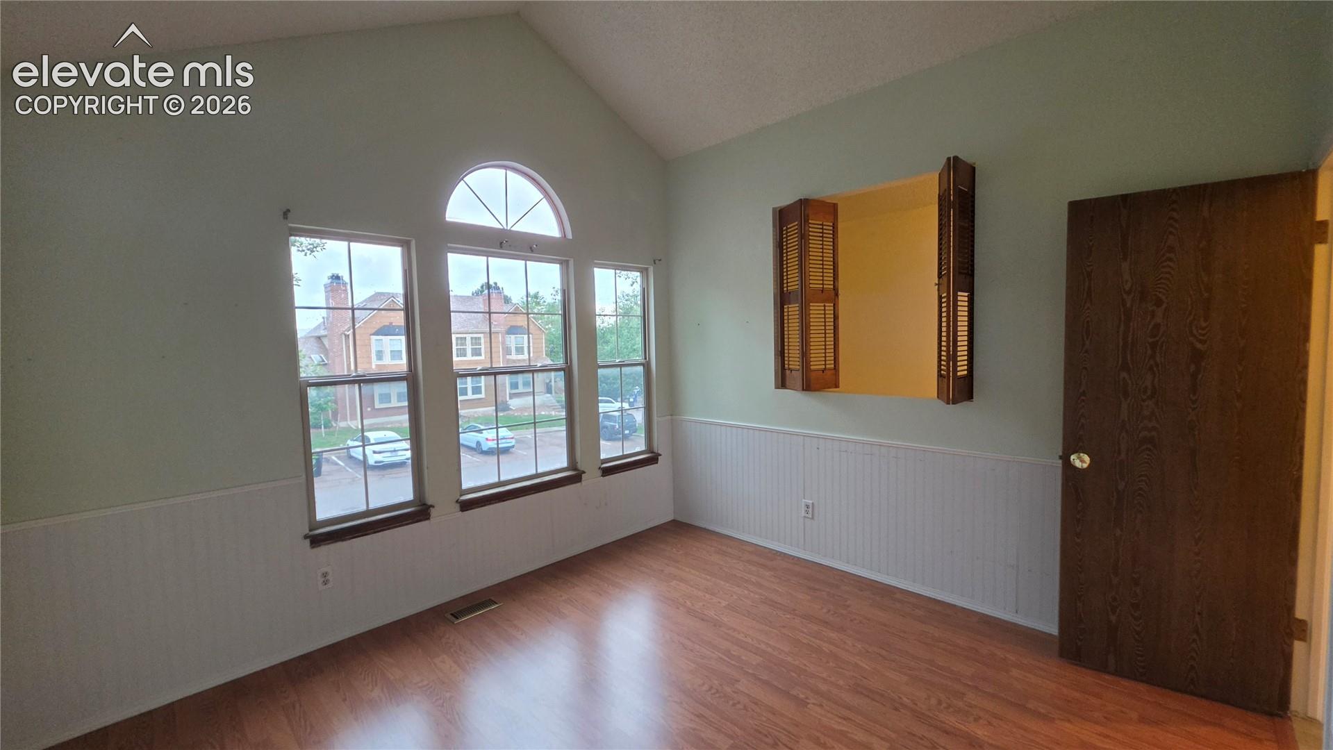 Vaulted ceiling with a large arched window, two vertical windows, wood-finish flooring, and white wainscoting