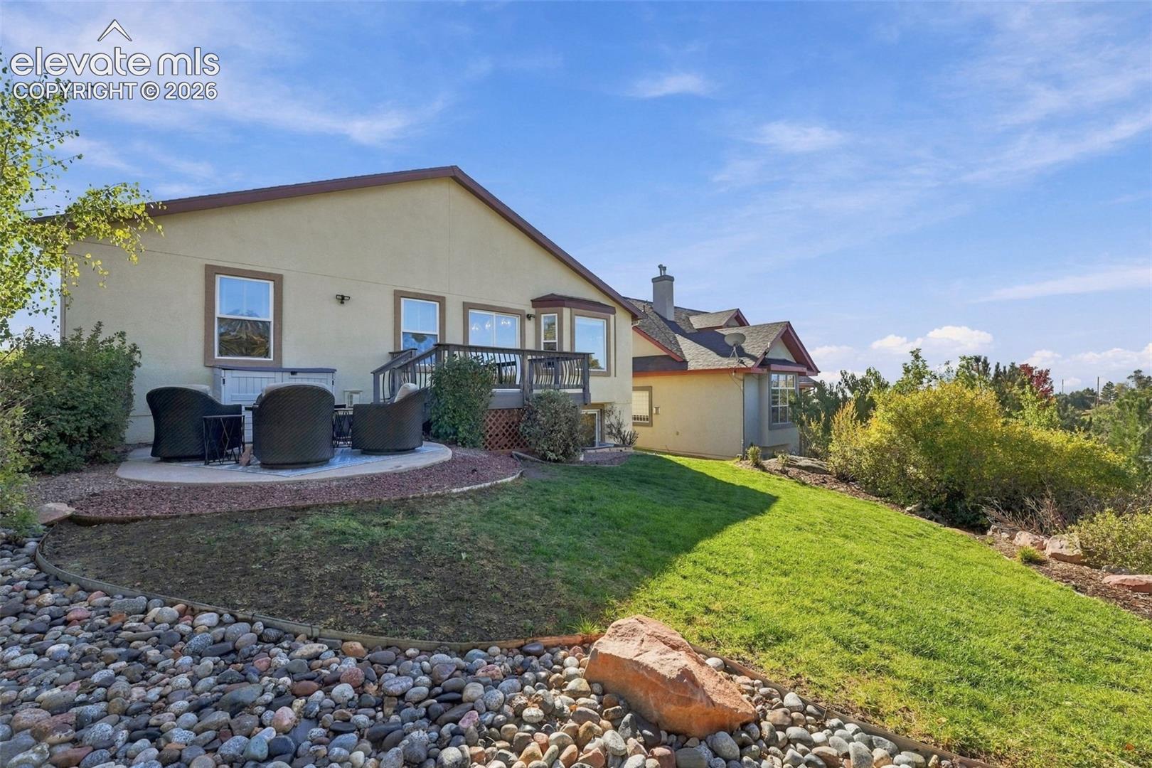 Stucco exterior featuring a raised wood deck, stone-bordered patio, terraced lawn, and rock garden landscaping