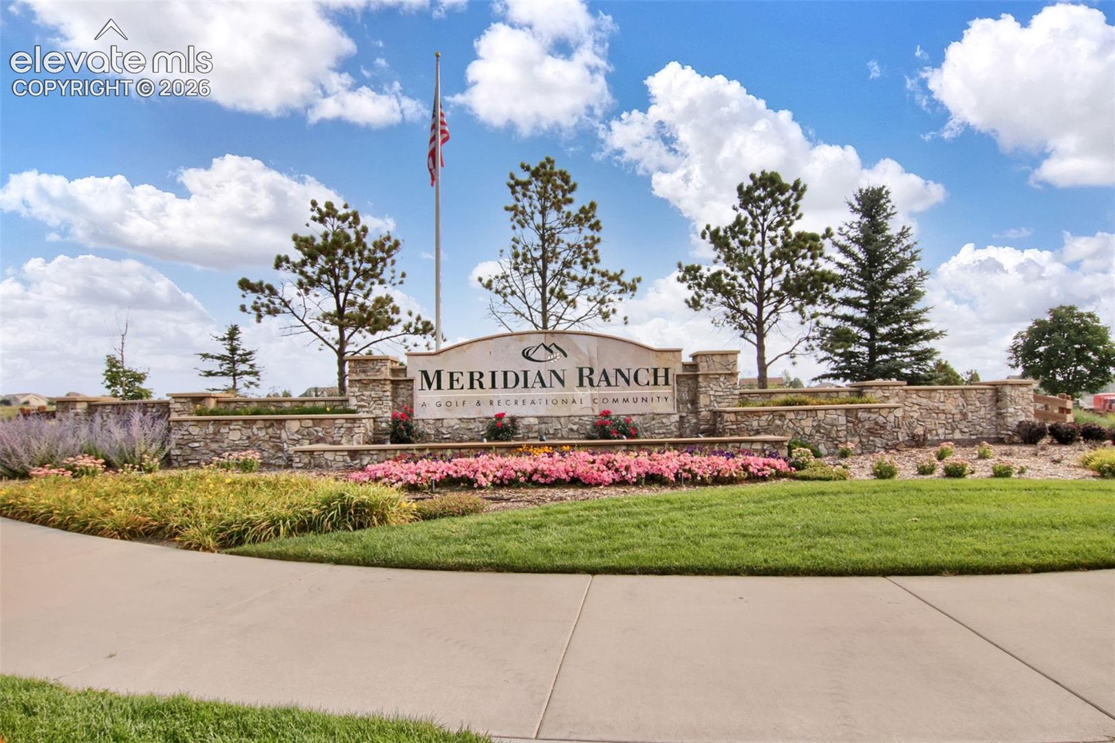 Stone-clad community entrance sign with integrated landscaping featuring flower beds, mature evergreen trees, and manicured lawn