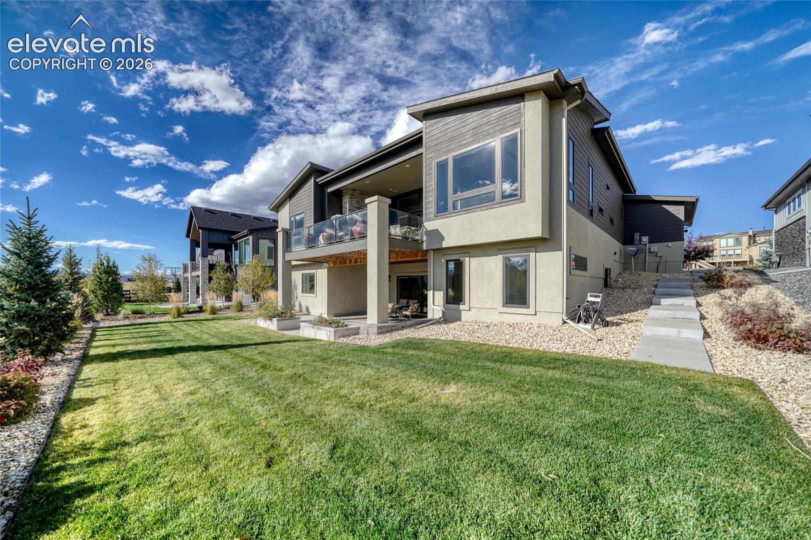 Rear exterior featuring a multi-level structure with mixed siding, a second-story balcony with glass railing, a covered ground-level patio, and a landscaped lawn
