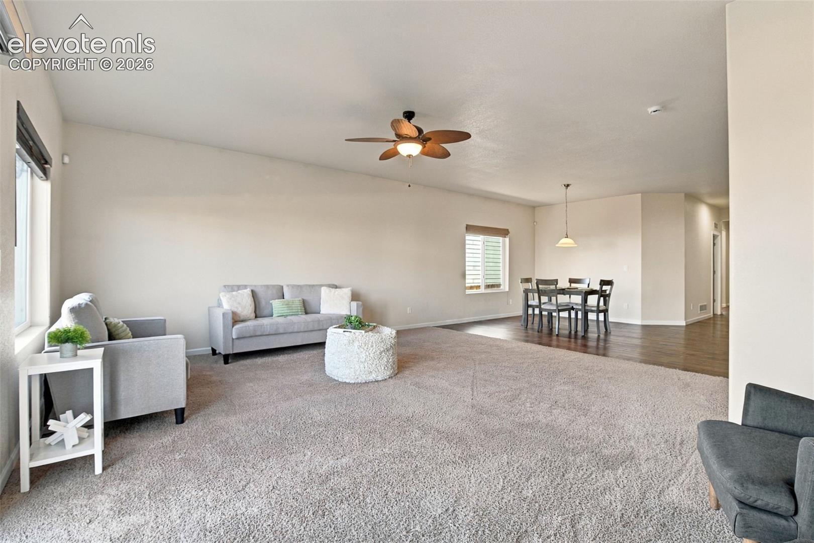 Open living area featuring neutral wall tones, multi-blade ceiling fan, plush carpeting, wood-finish flooring, and a pendant light fixture