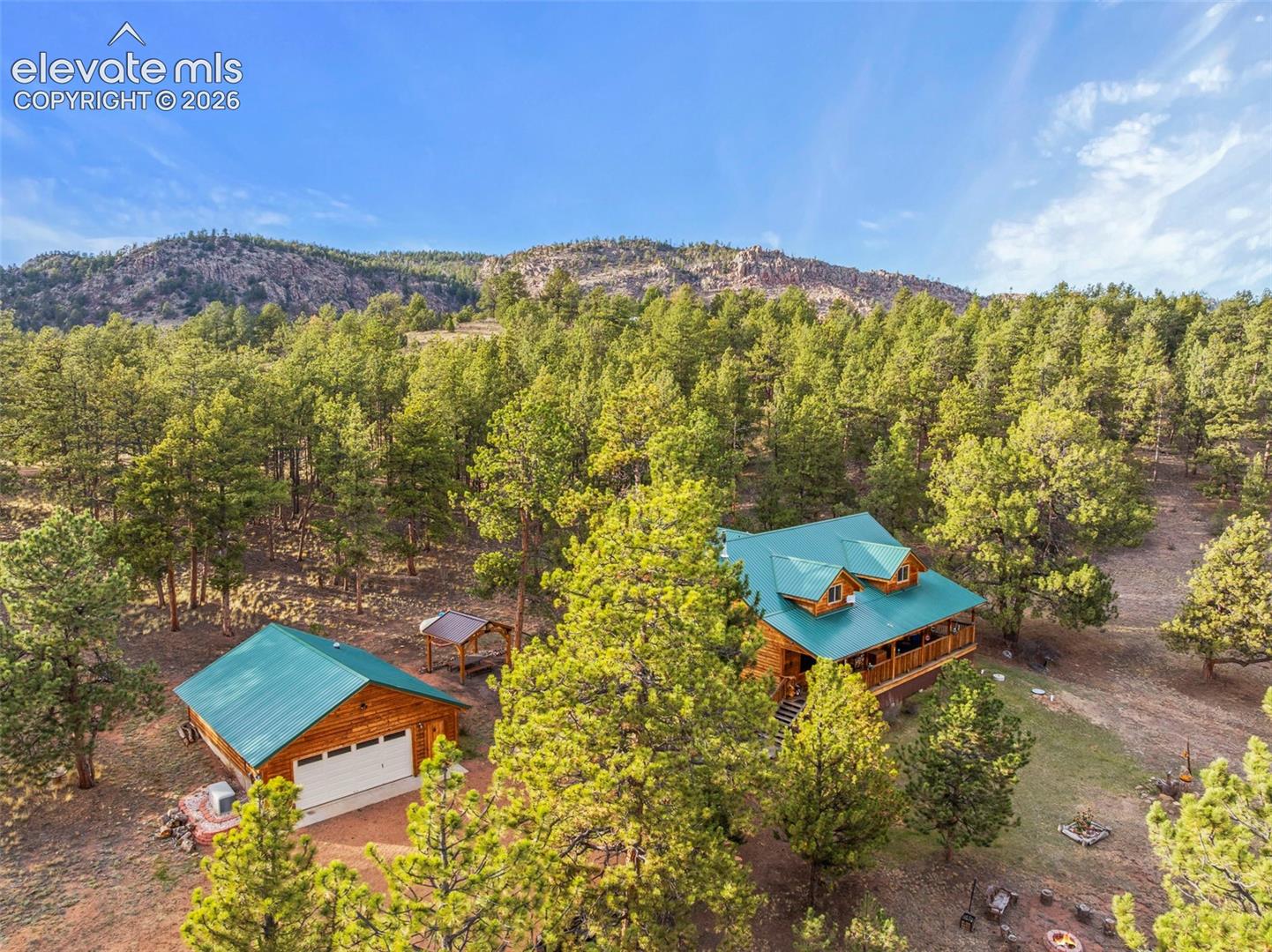 Log cabin exterior featuring a metal roof, expansive wrap-around deck, and detached garage, situated on a heavily wooded lot with mountain terrain
