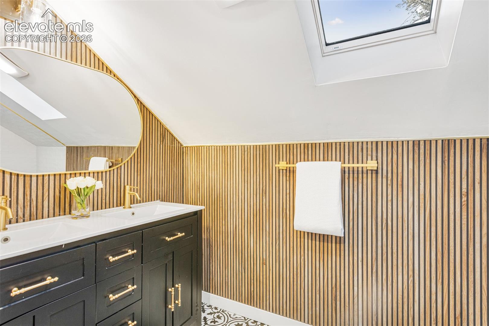 Bathroom featuring a dual-sink vanity with black cabinetry and gold-tone hardware, a gold-framed oval mirror, a vertical wood slat accent wall, and a skylight