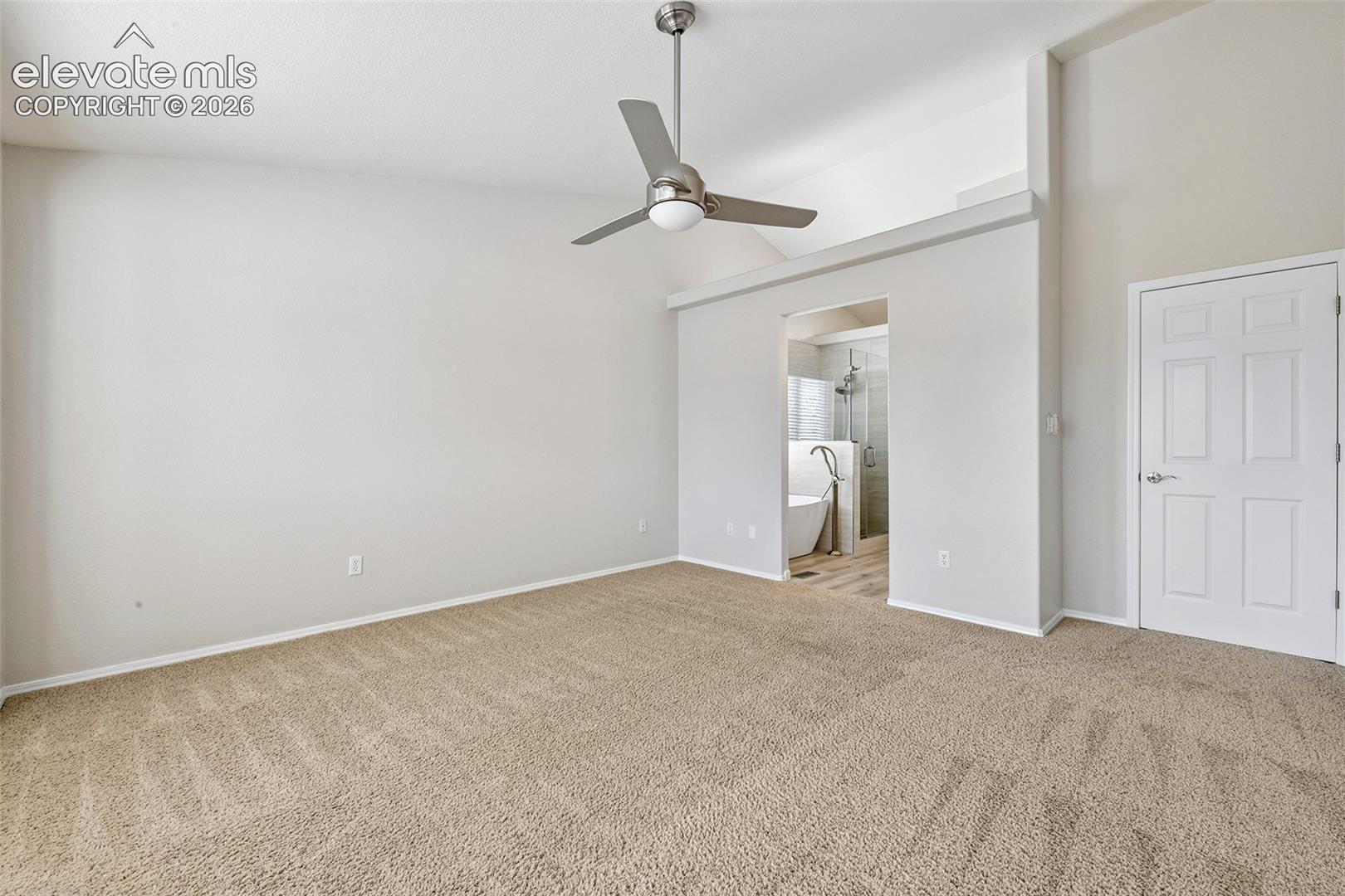 Vaulted ceiling with an attached ceiling fan, carpet flooring, interior doorway, and an open entryway leading to a bathroom