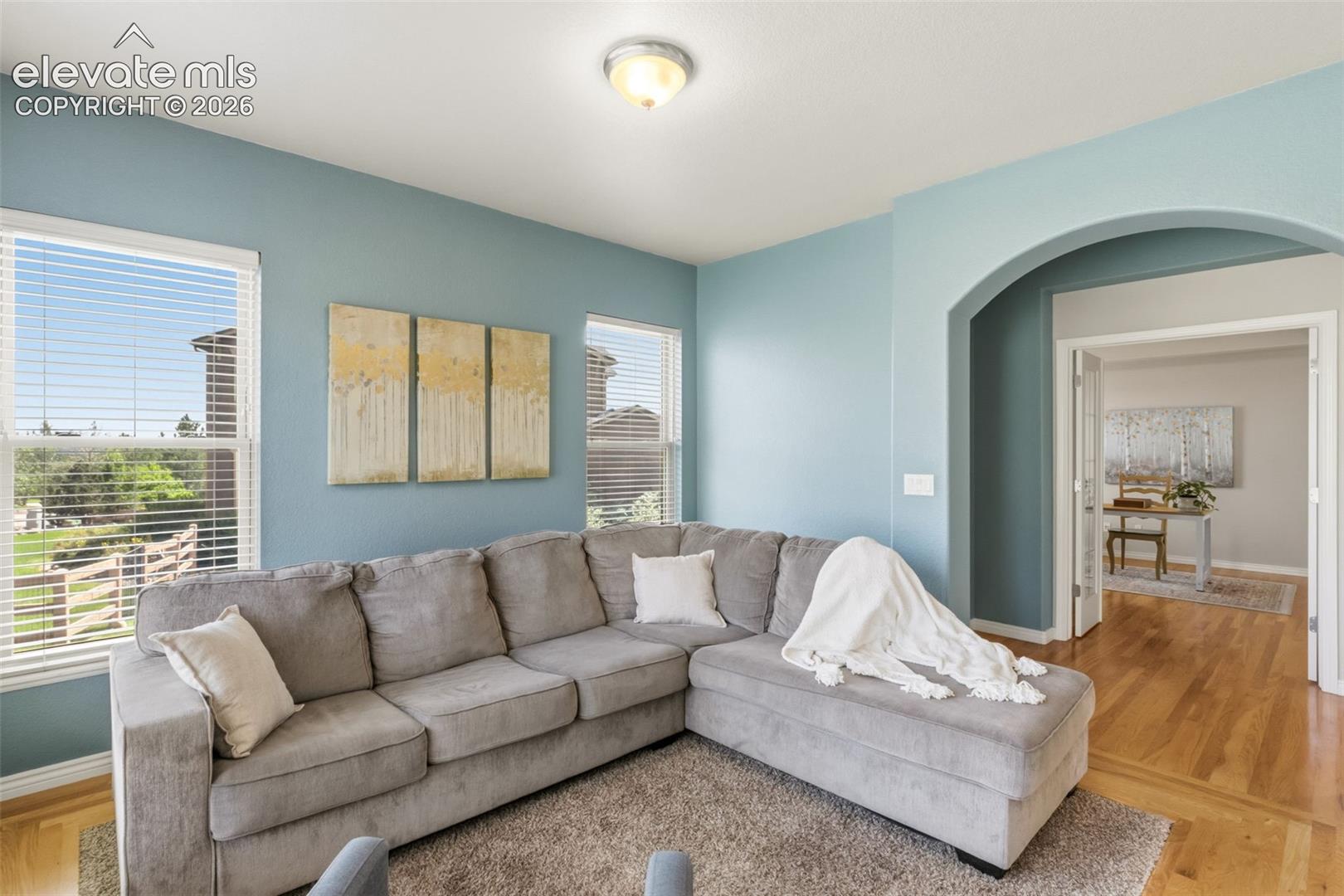 Living space featuring light blue wall paint, multiple windows with horizontal blinds, an arched doorway, and wood-finish flooring