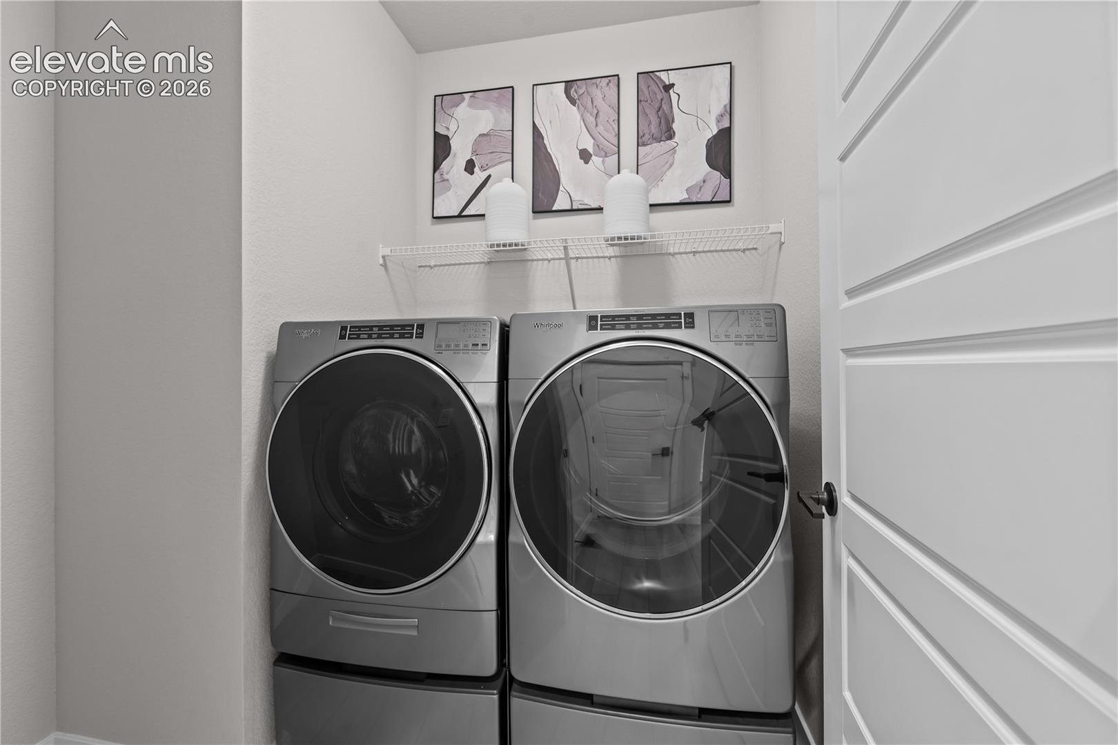 Laundry area featuring high-capacity stainless steel front-loading appliances, white wire shelving, and a paneled door with matte black hardware