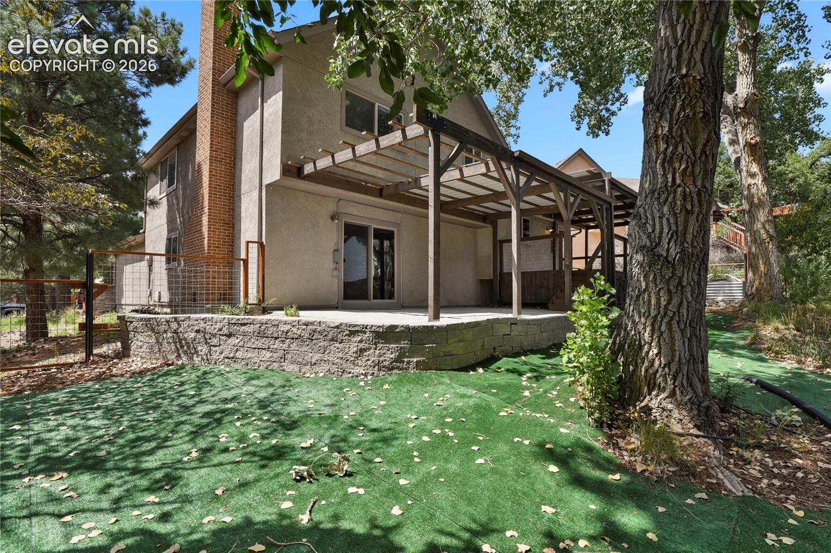 Rear exterior featuring a tiered stone retaining wall, covered pergola, brick chimney, and sliding glass door access