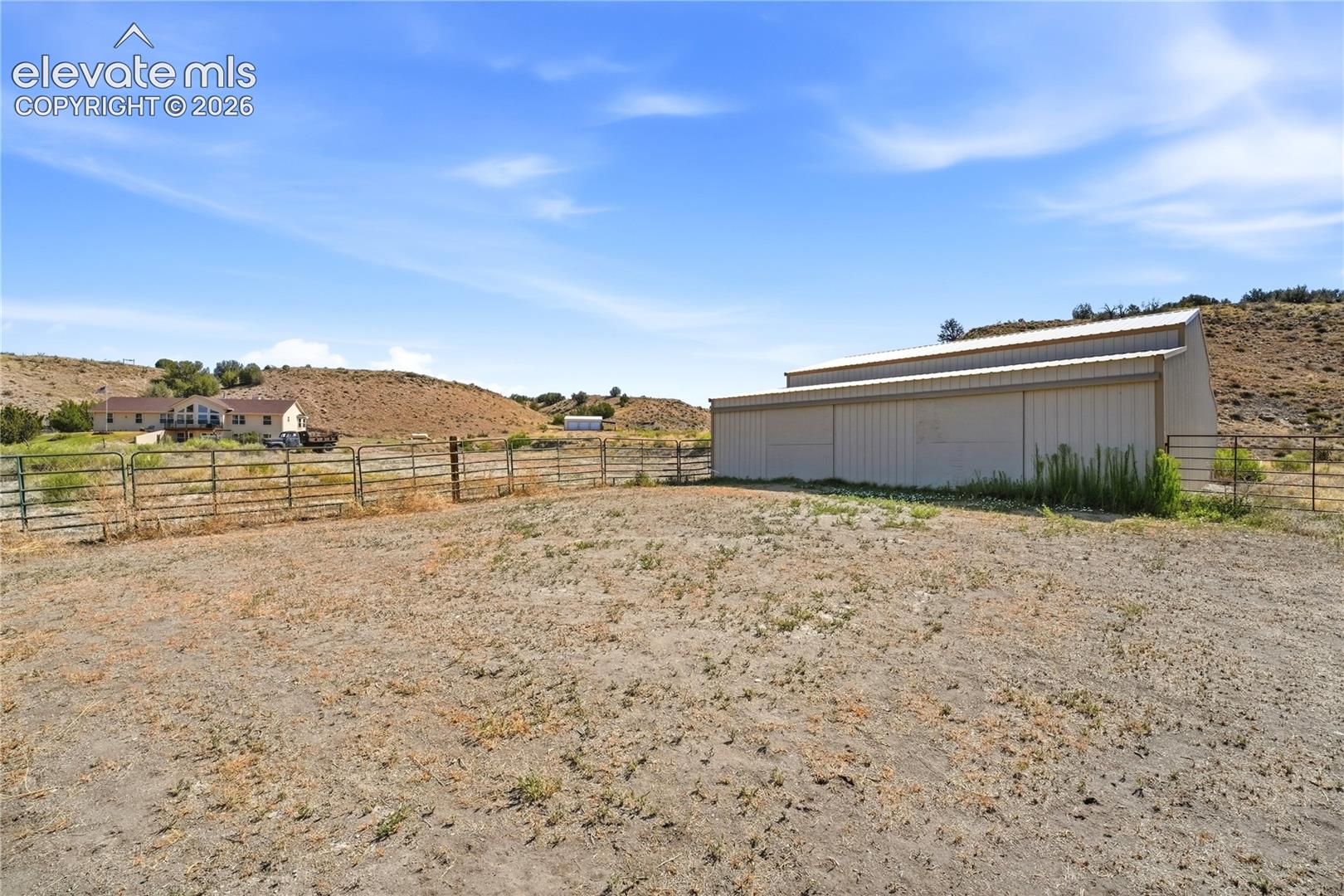 Expansive property featuring a large metal-sided outbuilding with a metal roof, perimeter fencing, and a distant residence with a deck