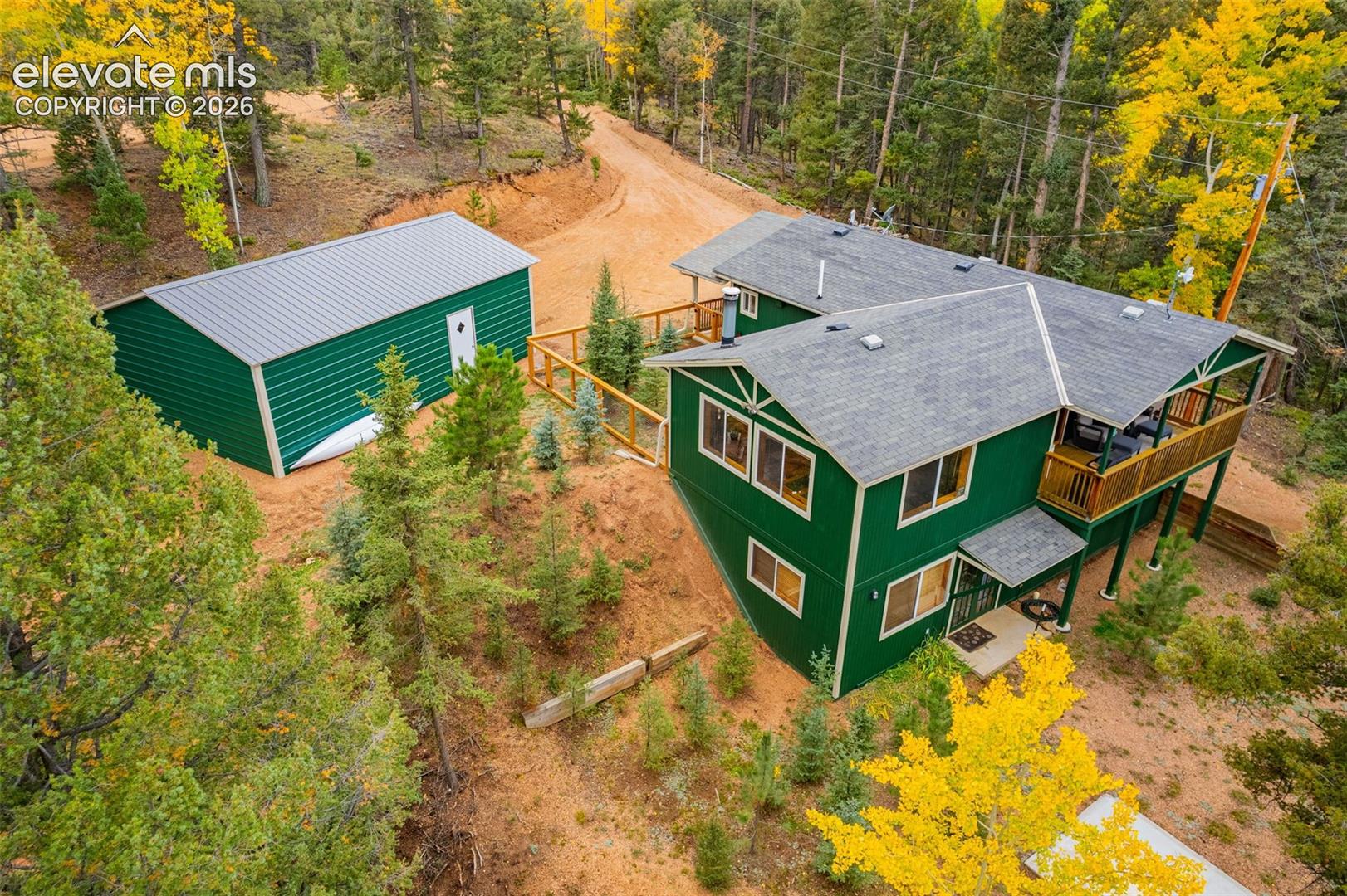 Two-story residence with green siding and shingle roofing, featuring an elevated wood deck, detached metal-sided storage building, and terraced landscape with mature evergreen and deciduous trees