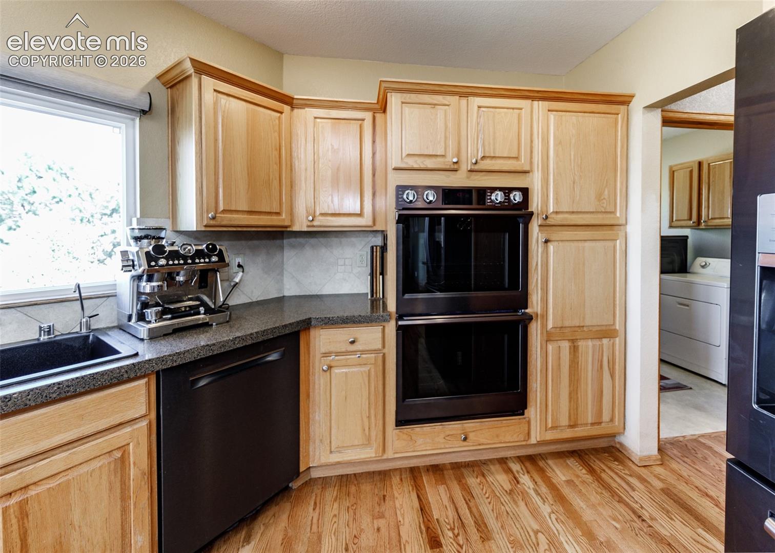 Wood-finish flooring, light wood cabinetry, double wall ovens, integrated dishwasher, and dark countertop with tile backsplash