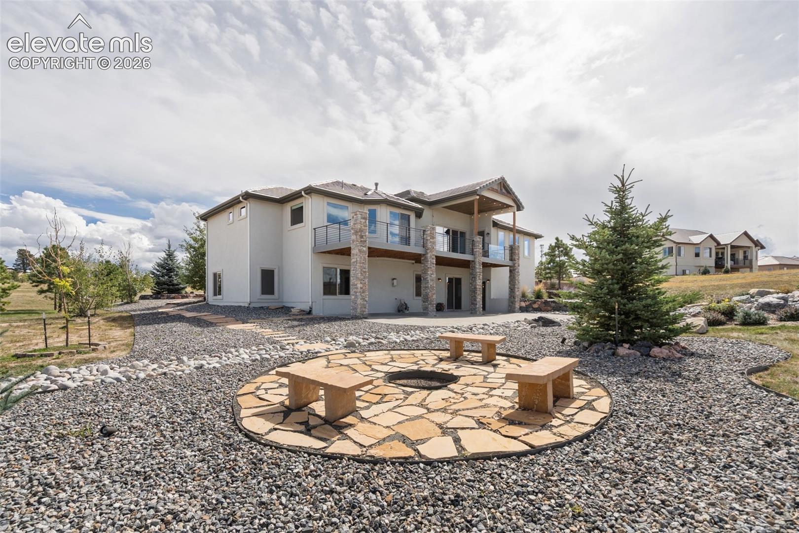 Expansive backyard featuring a stone patio with built-in fire pit, multiple balconies with glass railings, a gravel pathway, and mature trees