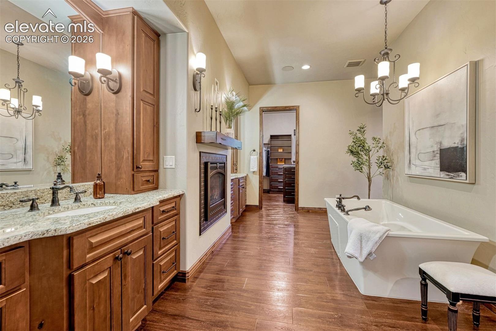 Wood-finish flooring leading to a freestanding soaking tub, dual vanity with a granite countertop, and an integrated fireplace