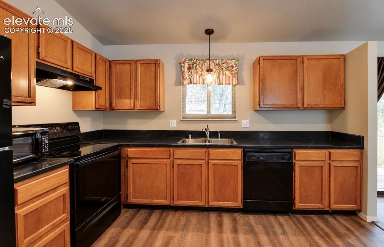 Kitchen featuring warm-tone wood cabinetry, dark solid-surface countertops, black integrated appliances, a dual-basin stainless steel sink, and wood-finish flooring