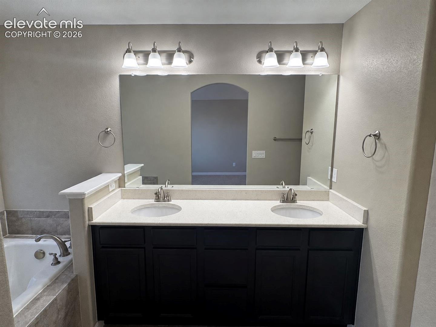 Double vanity featuring dark cabinetry, solid-surface countertops with dual undermount sinks, a large wall-mounted mirror, and twin three-light fixtures