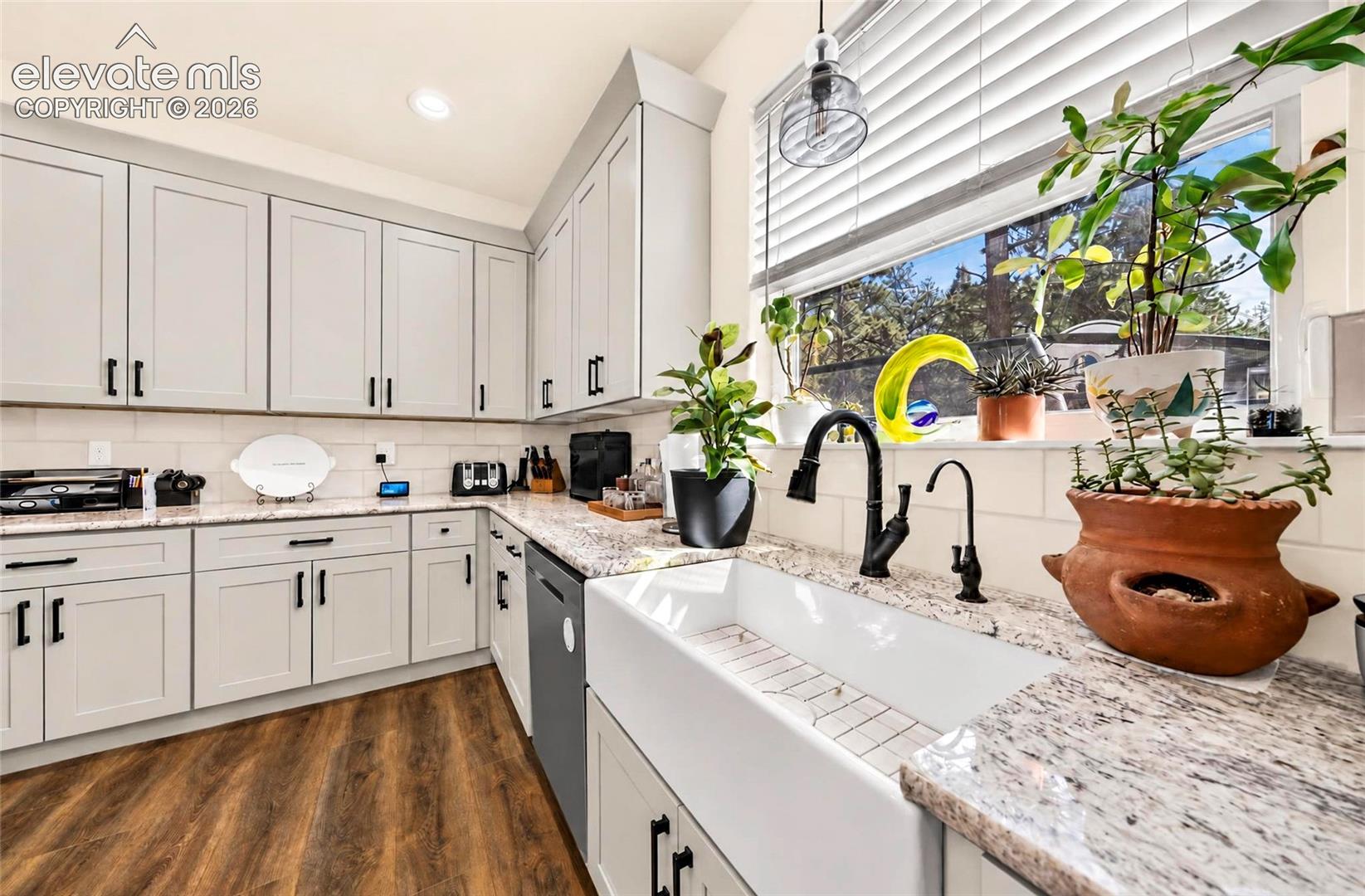 Kitchen featuring light gray cabinetry, stone-surface countertops, white ceramic farmhouse sink with matte black dual-faucet fixtures, light-toned subway tile backsplash, and wood-finish flooring