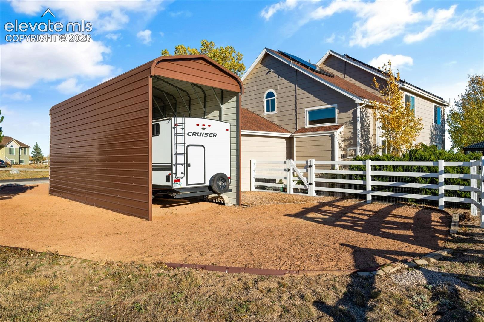Detached metal carport, graveled exterior parking area, and white post-and-rail perimeter fencing