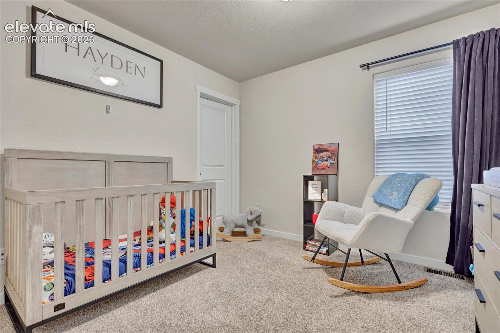 Bright nursery featuring neutral carpet flooring, a wooden crib with coordinating bedding, a cushioned glider chair, large windows with thermal blinds, and ample natural light throughout the space