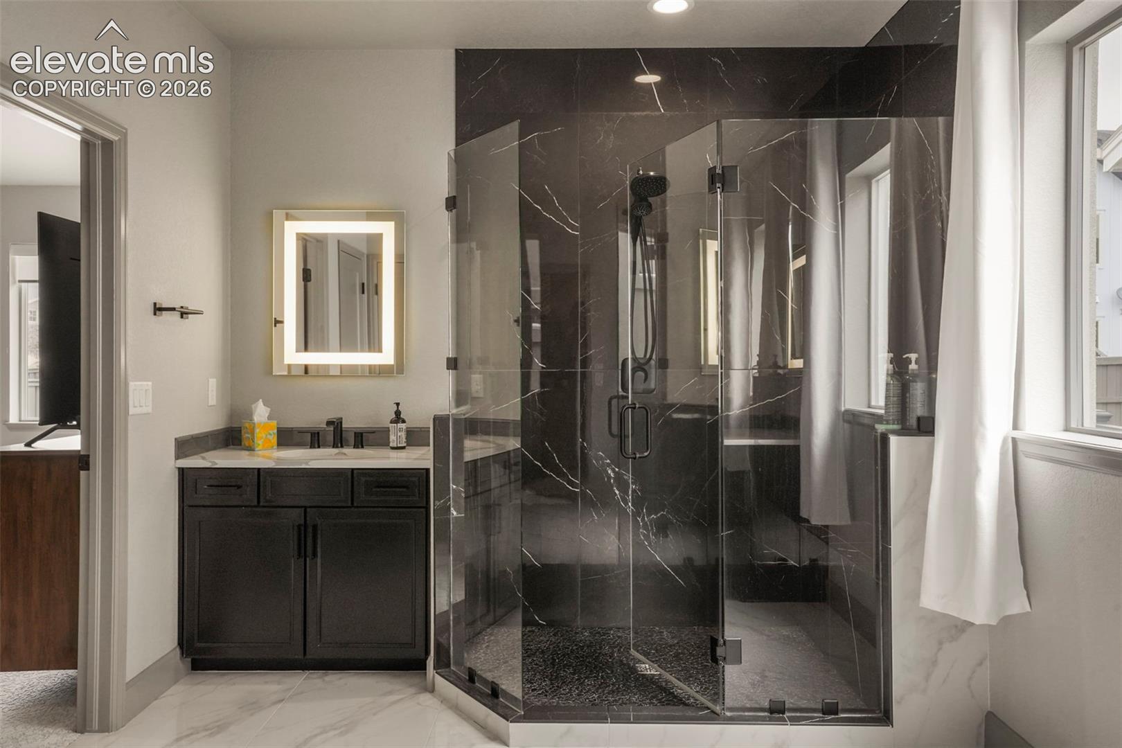 Spacious bathroom featuring a glass-enclosed shower with dark marble-style tile, a window, a single vanity with dark cabinetry, and an illuminated mirror