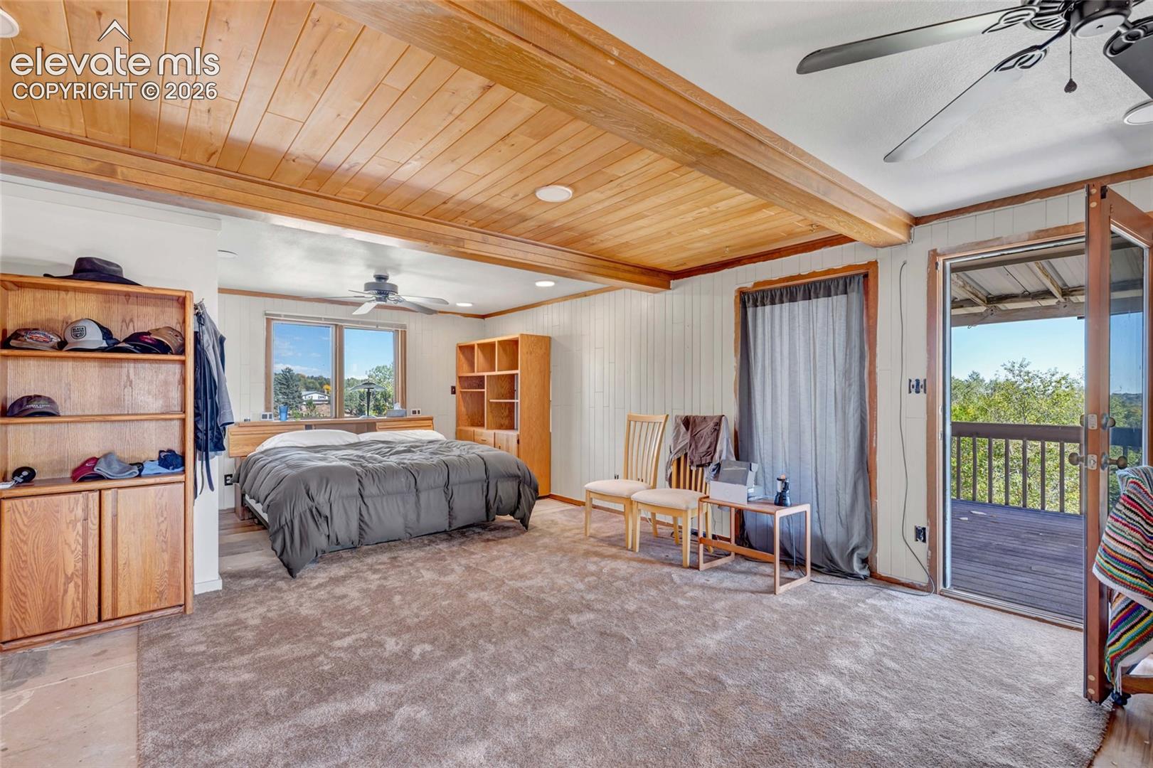 Wood-finish ceiling with exposed beams, light-colored carpeting, French doors opening to a deck, and built-in shelving
