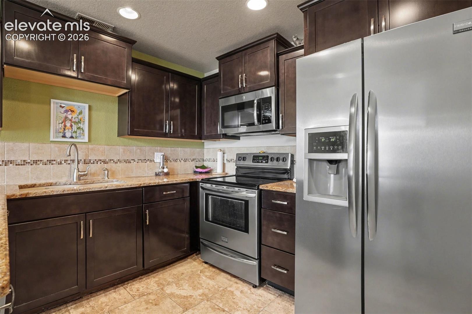 Kitchen featuring dark wood cabinetry, stainless steel appliances, stone-finish countertops, tiled backsplash with decorative inlay, and tile flooring