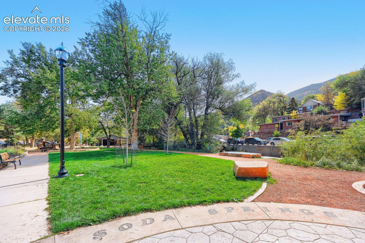 Manicured lawn area with mature trees, stone-look paver path, and a mountain backdrop